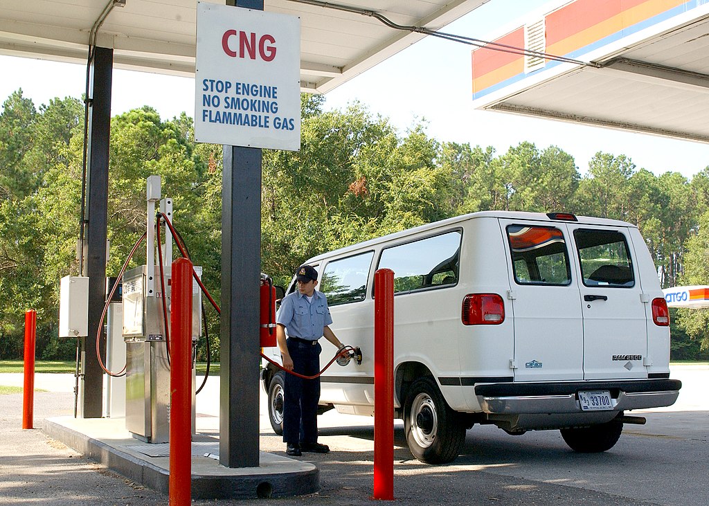 A man in the uniform of the United States navy refuels a government vehicle (a white van) at a compressed natural gas fuelling station on the naval base in Jacksonville, Florida. He stands directly below the sign saying "CNG. Stop engine. Stop smoking. Flammable gas." It is a sunny day.