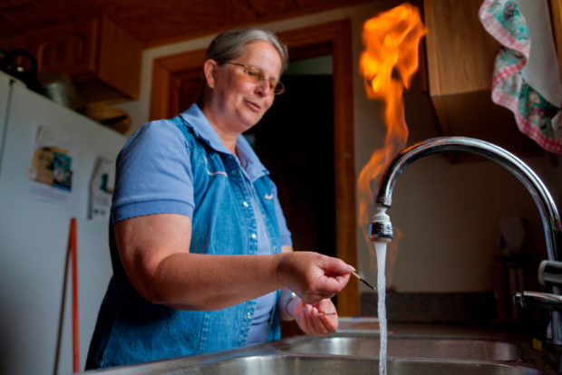 Woman sets fire to the water coming out of her kitchen tap. The water looks somewhat milky and she holds a match to it, now spent. An orange flame shoots up from the water. 