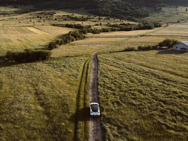 Aerial photograph of an electric vehicle driving on a one-track road with fields either side and a farmhouse or barn in the distance.