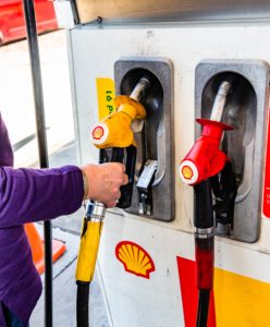 Close-up of petrol pumps dispending Shell fuel. A white person's right arm in a purple sleeve is holding the pump on the left.