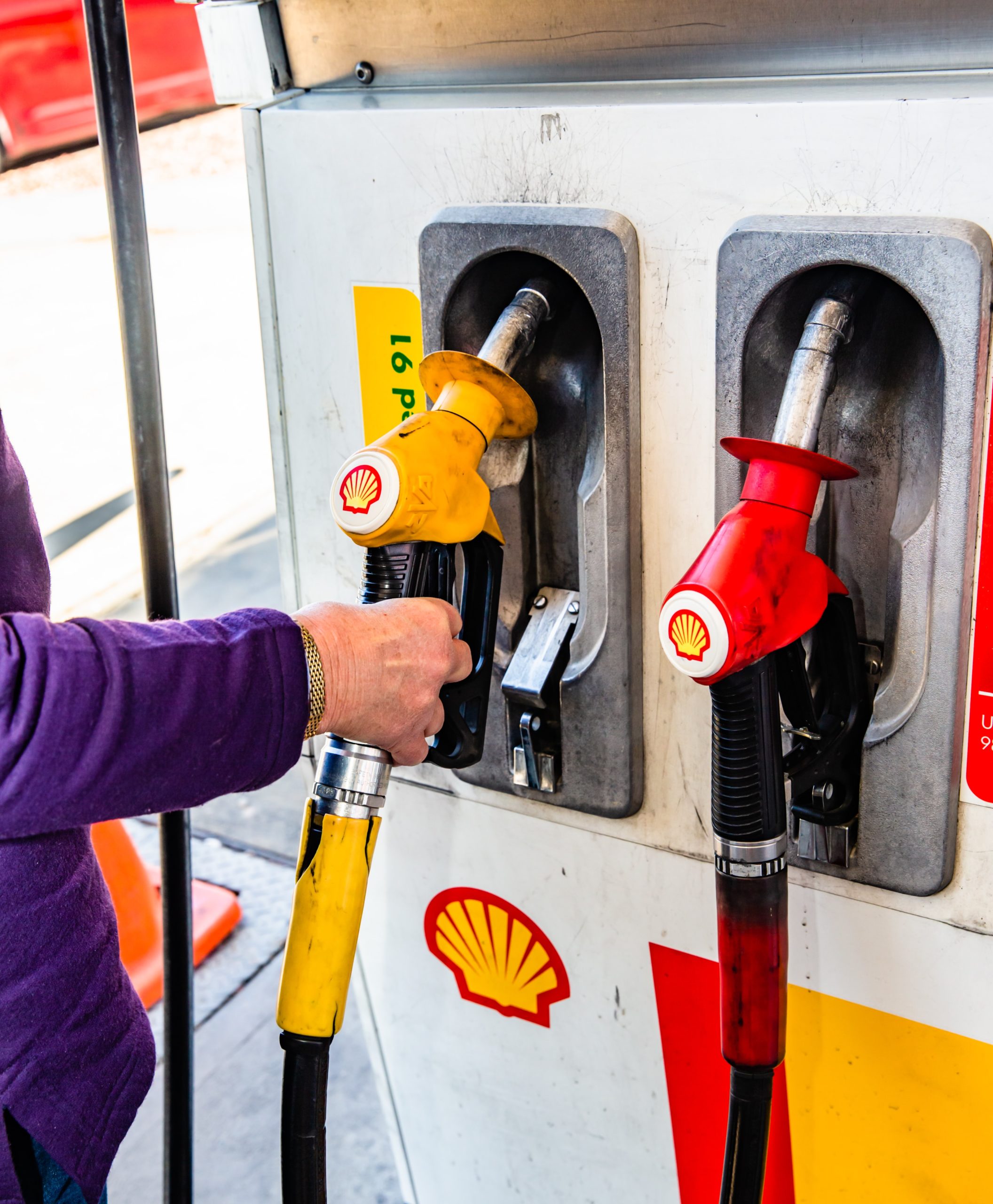 Close-up of petrol pumps dispending Shell fuel. A white person's right arm in a purple sleeve is holding the pump on the left.