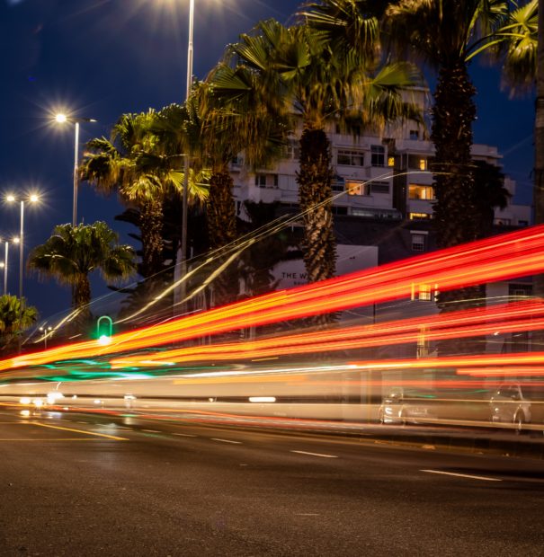 Photo of a Cape Town road at night. It is lined with palm trees and the lights of moving cars are blurred.