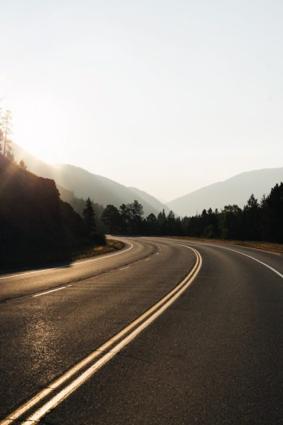 Photograph of a road curving into the distance with mountains on the left and forest on the right. More mountains in the distance. Picture probably taken at sunrise. 