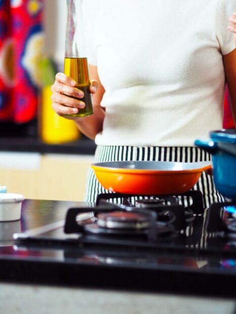 A person wearing a white top and striped black and white trousers stands by a stove with an unlabelled bottle of cooking oil in their right hand. There is an orange pan on the stove that they are probably about to pour the oil into.