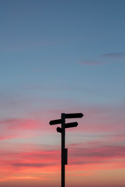 Photograph of a signpost at sunset. It is dark against the soft pinks of the sky so you can't read any of the locations on the sign.