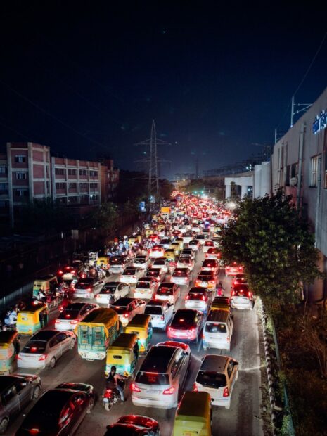 Overhead view of heavy traffic in Delhi. Vehicles are travelling six or seven abreast on a road with no lane markings. It is night and the cars have their lights on.